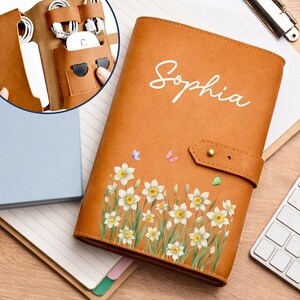 May include: A tan leather-like organizer case featuring the name Sophia in white script, decorated with white daffodil flowers and small butterflies. The case includes a snap closure and an interior section with pockets for cables and a mouse.