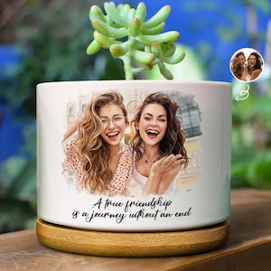 May include: A white cylindrical ceramic planter with a bamboo base, featuring a watercolour-style illustration of two smiling people and the printed text: A true friendship is a journey without an end. A green succulent plant grows from the top.