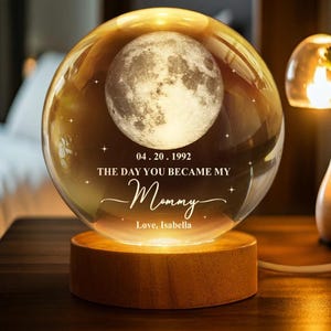 Puede incluir: Una esfera de cristal iluminada con una imagen detallada de la luna, sobre una base de madera. La esfera tiene el texto "04.20.1992 THE DAY YOU BECAME MY Mommy Love, Isabella". La luz cálida crea un ambiente acogedor.
