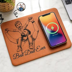 Op de afbeelding: Een bruin draadloos oplaadpad met een zwart-wit illustratie van een vader en kind. De woorden "Best Dad Ever" zijn cursief geschreven. Een smartphone wordt op het pad opgeladen.