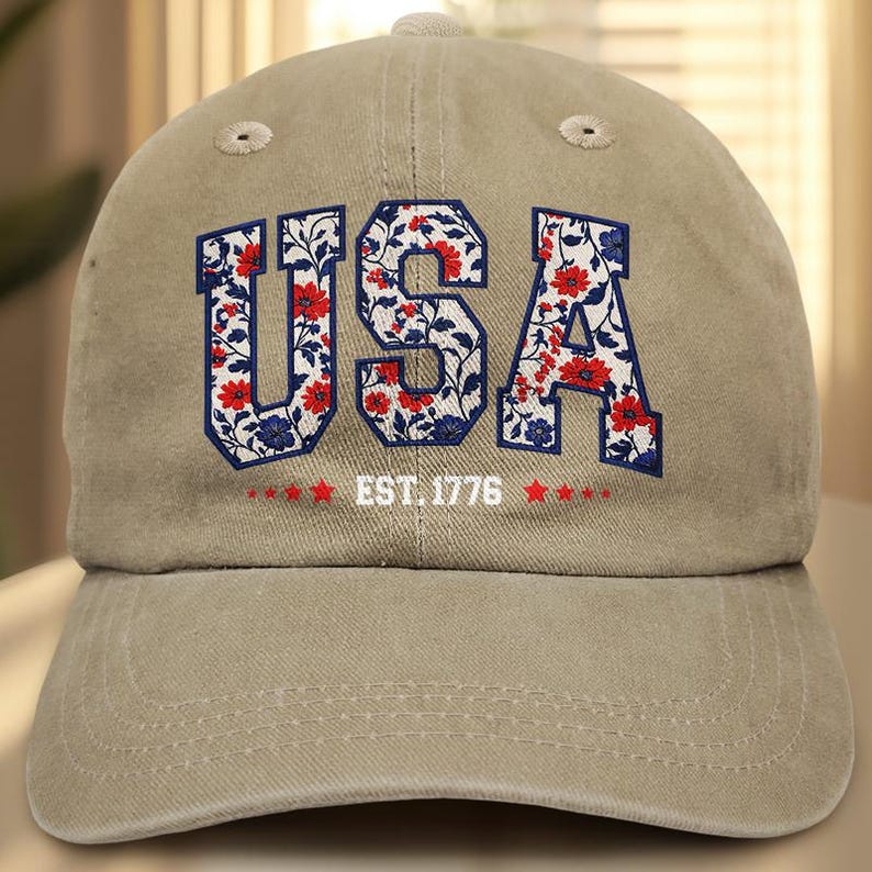 Puede incluir: Gorra de b&eacute;isbol beige con la palabra "USA" en letras florales. Las letras est&aacute;n rellenas de flores rojas y blancas, delineadas en azul. El texto "EST. 1776" est&aacute; debajo, con estrellas rojas a cada lado.