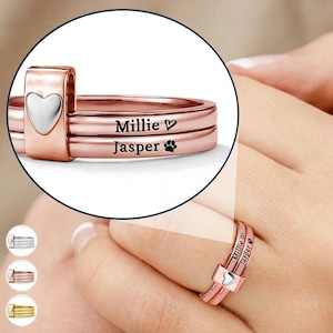 May include: Rose gold rings with the names "Millie" and "Jasper" engraved on them. One ring has a heart-shaped accent, and another has a paw print. The rings are stacked together, and one is worn on a finger.