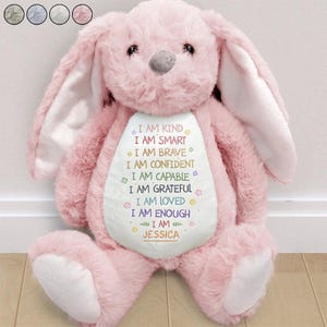 May include: A large, plush pink bunny rabbit with long ears and a white belly. The belly features colorful text that reads "I AM KIND, I AM SMART, I AM BRAVE, I AM CONFIDENT, I AM CAPABLE, I AM GRATEFUL, I AM LOVED, I AM ENOUGH" and the name "JESSICA".
