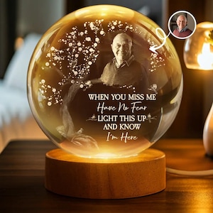 Puede incluir: Una esfera de cristal brillante con la foto de un hombre y el texto "WHEN YOU MISS ME Have No Fear LIGHT THIS UP AND KNOW I'm Here". La esfera se asienta sobre una base de madera, iluminada desde dentro, creando un brillo cálido.