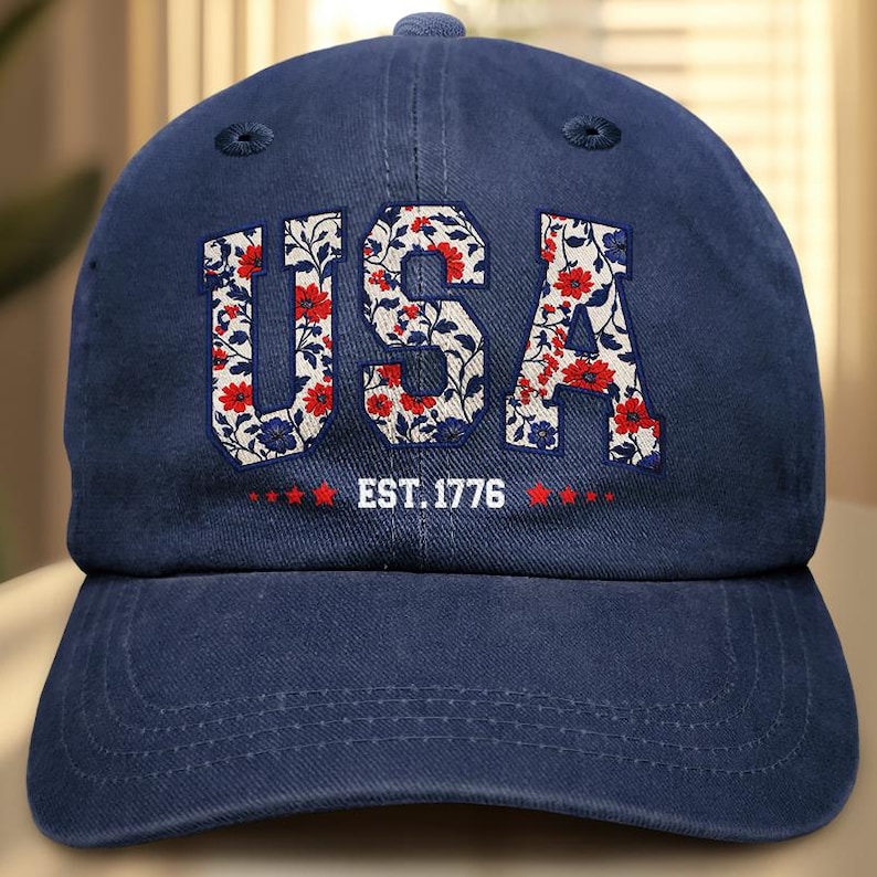 Puede incluir: Gorra de b&eacute;isbol azul marino con la palabra "USA" en letras blancas rellenas de un estampado floral de flores rojas y hojas azules. El texto "EST. 1776" est&aacute; debajo.