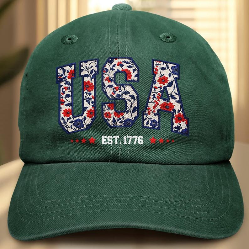 Puede incluir: Gorra de b&eacute;isbol verde oscuro con la palabra "USA" en letras blancas rellenas de un estampado floral de flores rojas y azules. El texto "EST. 1776" est&aacute; debajo de la palabra "USA".