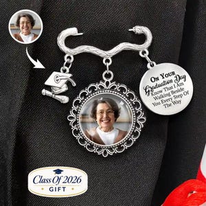 May include: A silver-toned graduation brooch with a photo pendant and a mortarboard charm. The pendant has a photo of a woman and the text "On Your Graduation Day Know That I Am Walking Beside You Every Step Of The Way". The brooch also has the text "Class Of 2026 GIFT".