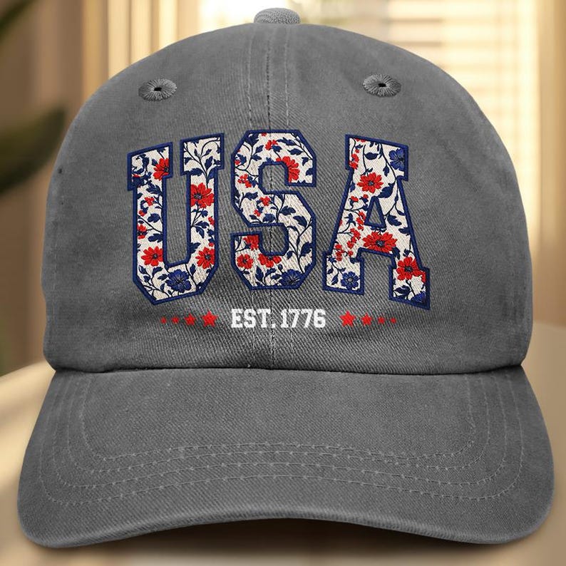 Puede incluir: Gorra de b&eacute;isbol gris con la palabra "USA" en un estampado floral de flores rojas y azules. El texto "EST. 1776" est&aacute; debajo. La gorra tiene una visera curva y un bot&oacute;n en la parte superior.