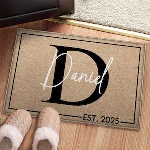 Puede incluir: Una alfombra rectangular con fondo beige y borde negro. La alfombra presenta una gran letra negra "D" con el nombre "Daniel" en escritura blanca. Debajo del nombre está "EST. 2025" en negro.