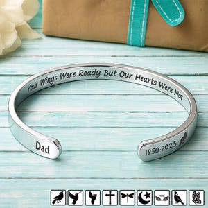 Op de afbeelding: Zilverkleurige manchetarmband met de inscriptie "Your Wings Were Ready But Our Hearts Were Not". De armband heeft ook het woord "Dad" en de jaren "1950-2025". Een gedenkgeschenk.
