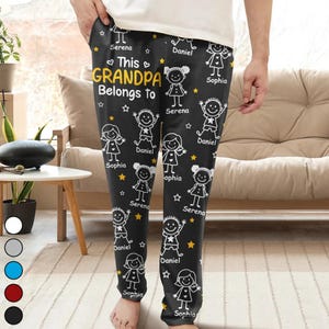 Könnte beinhalten: Schwarze Schlafanzughose mit einem sich wiederholenden Muster aus Cartoon-Kinderzeichnungen, Sternen und den Namen "Serena", "Daniel" und "Sophia". Der Text "This GRANDPA Belongs To" ist gelb. Die Hose wird von einer Person getragen.