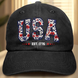Puede incluir: Gorra de b&eacute;isbol negra con la palabra "USA" en letras blancas rellenas de un estampado floral de flores rojas y azules. El texto "EST. 1776" est&aacute; debajo de la palabra "USA".