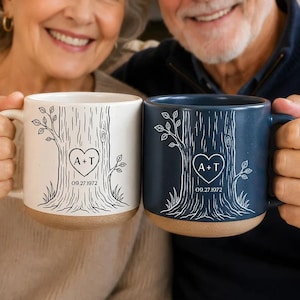 May include: Two ceramic mugs, one cream and one navy blue, each featuring a white or black line-art illustration of a tree trunk with a heart carved into the center. Inside the heart, the letters A + T are printed above the date 09.27.1972.