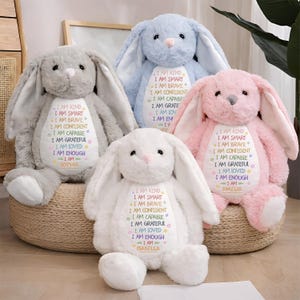 May include: Five soft toy bunny rabbits in pastel shades of grey, blue, pink, and white. Each has long ears and embroidered text on its chest, including phrases like "I AM KIND" and personalized names. They are arranged on a woven basket.