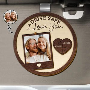 May include: A circular wooden car visor clip featuring the text "DRIVE SAFE I Love You" at the top. It includes a rectangular photo frame with a picture of a couple, a small heart cutout, and a heart-shaped puzzle piece with the names "WILLIAM" and "JENNIFER" engraved on it.