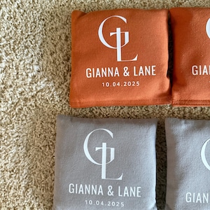 Könnte beinhalten: Vier quadratische Beanbags, zwei in Orange und zwei in Grau, sind auf einer strukturierten Oberfläche angeordnet. Jeder Beutel zeigt die weißen Initialen "GL" und die Namen "GIANNA & LANE" mit dem Datum "10.04.2025" darunter.