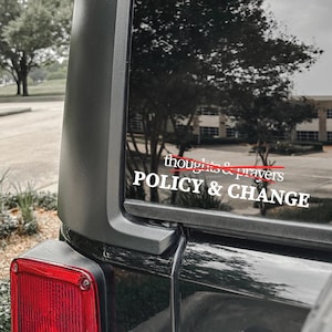 May include: A black vehicle's rear window displays a white decal. The decal reads "thoughts & prayers" with a red line through it, and below it, "POLICY & CHANGE" in bold white letters. The background shows trees and a building.