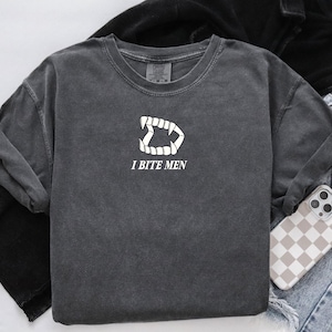 May include: Dark gray crew neck sweatshirt with a white graphic of a set of teeth and the text "I BITE MEN". The sweatshirt is styled with a black jacket, jeans, and a phone case with a checkered pattern.