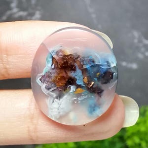 May include: A clear, oval-shaped resin pendant with an internal landscape of brown, blue, and white formations. The pendant has a polished surface and appears to be handmade, showcasing a unique, artistic design. The colors and textures create a miniature, abstract scene.