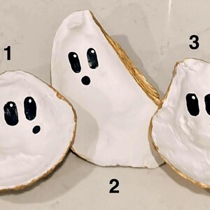 Peut inclure: Trois coquilles d'huîtres blanches peintes avec des visages de fantômes de dessins animés. Chaque coquille a des yeux ovales noirs et un point noir pour la bouche. Les bords des coquilles sont peints en or. Les coquilles sont disposées sur une surface gris clair.