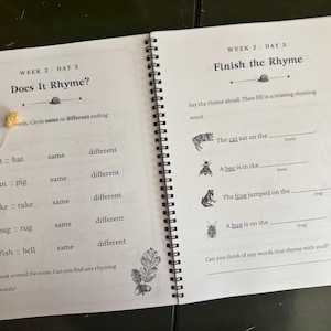 May include: A spiral-bound educational workbook open to two pages. The left page is titled Week 2 Day 2 Does It Rhyme? with word pairs to compare. The right page is titled Week 2 Day 3 Finish the Rhyme with fill-in-the-blank sentences and illustrations.