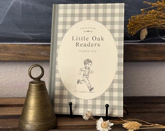 May include: A hardcover book titled Little Oak Readers Primer One with a light gray and white gingham cover pattern. The cover features a central oval illustration of a child running. Beside the book sits a weathered brass bell with a ring handle.