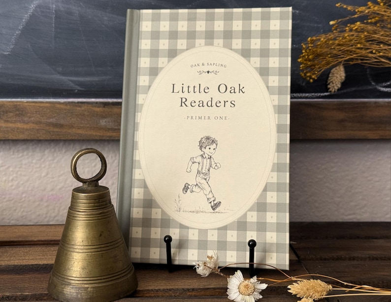 Puede incluir: Un libro de tapa dura titulado Little Oak Readers Primer One con un patr&oacute;n de cuadros vichy en gris claro y blanco. La portada presenta una ilustraci&oacute;n ovalada central de un ni&ntilde;o corriendo. Junto al libro hay una campana de lat&oacute;n.