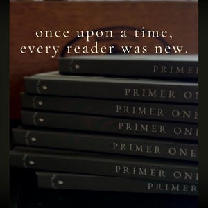 Puede incluir: Una pila de siete libros de color gris oscuro con el texto "PRIMER ONE" impreso en blanco en cada lomo. El texto "once upon a time, every reader was new." aparece en blanco sobre los libros. Cada lomo presenta un peque&ntilde;o icono de bellota.