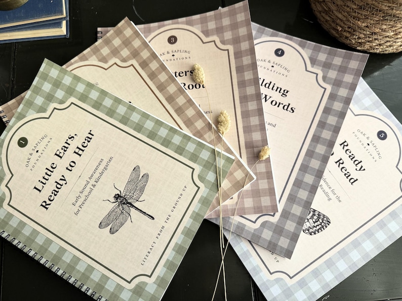 May include: A set of five spiral-bound educational workbooks with gingham patterned covers in sage green, tan, brown, gray, and light blue. Each cover features a decorative label with the text Oak & Sapling Foundations, numbered titles, and illustrations.