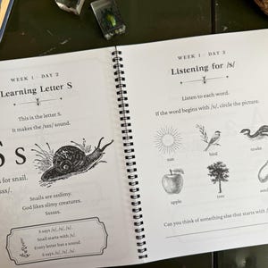 May include: A spiral-bound educational workbook open to two pages. The left page features the letter S, a snail illustration, and text about the /s/ sound. The right page titled Listening for /s/ displays illustrations of a sun, bird, snake, apple, tree, and seed.