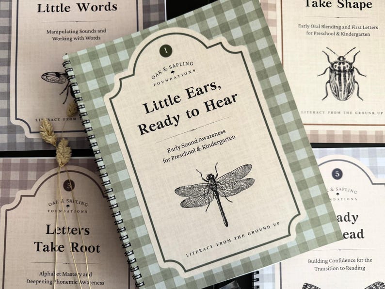 May include: A spiral-bound educational workbook titled Little Ears, Ready to Hear with a green and white gingham cover. The cover features a black line drawing of a dragonfly, the text Oak & Sapling Foundations, and Early Sound Awareness.