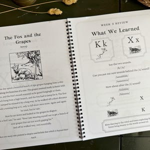 May include: A spiral-bound educational workbook open to two pages. The left page features the fable The Fox and the Grapes with an illustration. The right page is titled Week 3 Review What We Learned, focusing on the letters Kk and Xx with phonics exercises.