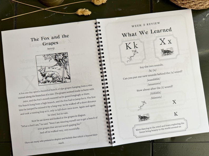 May include: A spiral-bound educational workbook open to two pages. The left page features the fable The Fox and the Grapes with an illustration. The right page is titled Week 3 Review What We Learned, focusing on the letters Kk and Xx with phonics exercises.