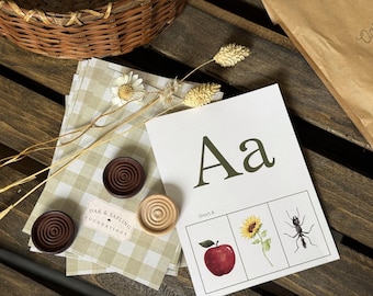 May include: A white educational card featuring the letter Aa in dark green, with the text Short A and illustrations of a red apple, a yellow sunflower, and a black ant. The card is accompanied by two dark brown and one light wooden circular tokens.