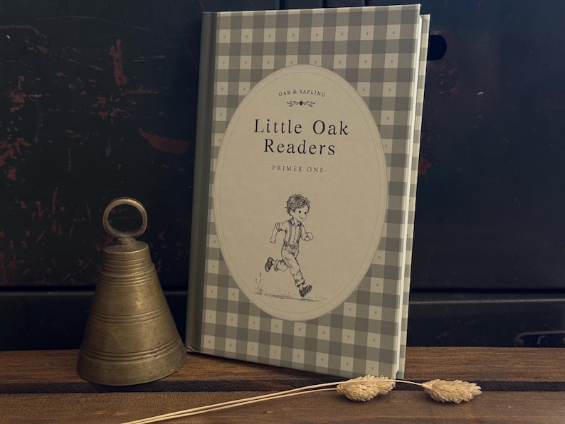 Puede incluir: Un libro de tapa dura titulado Little Oak Readers Primer One con un patr&oacute;n de cuadros vichy gris y blanco en la cubierta. La portada presenta un &oacute;valo central con un dibujo lineal de un ni&ntilde;o corriendo. Junto al libro hay una campana de lat&oacute;n y dos vainas secas.