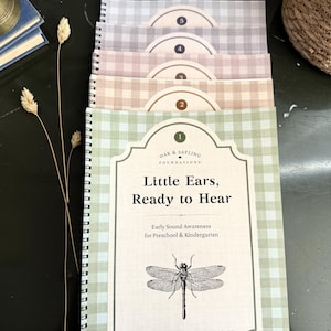 May include: A stack of five spiral-bound educational workbooks with gingham covers in pastel shades of green, tan, mauve, blue, and grey. Each cover features a numbered label, the text Oak & Sapling Foundations, Little Ears, Ready to Hear, and a dragonfly.