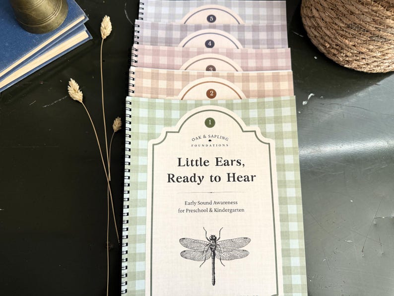 May include: A stack of five spiral-bound educational workbooks with gingham covers in pastel shades of green, tan, mauve, blue, and grey. Each cover features a numbered label, the text Oak & Sapling Foundations, Little Ears, Ready to Hear, and a dragonfly.