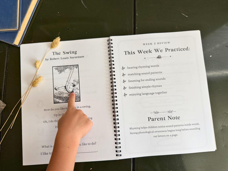 May include: An open spiral-bound educational workbook featuring the poem The Swing by Robert Louis Stevenson on the left page and a Week 2 Review section on the right page. The text includes practice goals like hearing rhyming words and a parent note.
