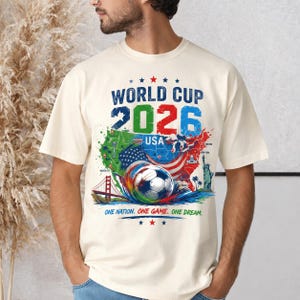 May include: Cream-colored t-shirt featuring a colorful graphic for the 2026 World Cup USA. The design includes a soccer ball, maps of the USA, and the text "World Cup 2026 USA". The shirt also has the text "One Nation. One Game. One Dream."