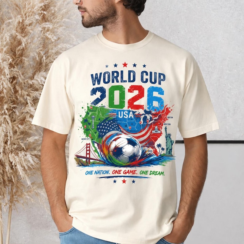 May include: Cream-colored t-shirt featuring a colorful graphic for the 2026 World Cup USA. The design includes a soccer ball, maps of the USA, and the text "World Cup 2026 USA". The shirt also has the text "One Nation. One Game. One Dream."