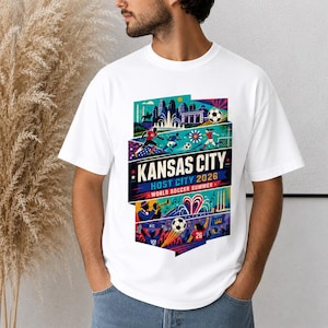 K&ouml;nnte beinhalten: Ein wei&szlig;es T-Shirt mit einem farbenfrohen Grafikdruck von Kansas City als Gastgeberstadt 2026. Das Design zeigt Fu&szlig;ballspieler, einen Fu&szlig;ball, eine Stadtansicht, einen Jazzmusiker und den Text KANSAS CITY HOST CITY 2026 WORLD SOCCER SUMMER.