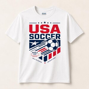May include: White t-shirt with a graphic design featuring "USA SOCCER" in red and blue. The design includes a soccer ball with speed lines, stars, and the text "WORLD CUP 2026". The shirt is made of a soft material.