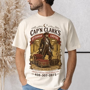 May include: A cream-coloured t-shirt featuring a graphic of a pirate standing in a hallway with a red armchair, ottoman, and lantern. The text reads: CAP'N CLARK'S OTTOMAN EMPIRE, FINE FURNITURE FROM THE BACKROOMS, LIMITLESS CORRIDORS, ENDLESS POSSIBILITIES, 408-307-2873.
