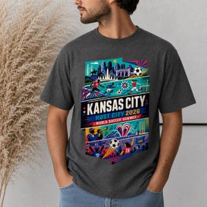 K&ouml;nnte beinhalten: Ein dunkelgraues T-Shirt mit einem farbenfrohen Grafikdruck und dem Text KANSAS CITY HOST CITY 2026 WORLD SOCCER SUMMER. Das Design zeigt Illustrationen von Fu&szlig;ballspielern, einer Stadtsilhouette, einem Trompeter und Fu&szlig;b&auml;llen.