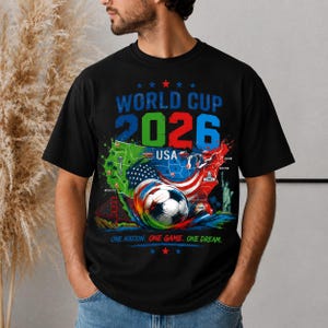 May include: Black t-shirt with a colorful graphic design for the 2026 World Cup in the USA. The design includes a soccer ball, the USA map, and the text "World Cup 2026 USA" and "One Nation. One Game. One Dream."