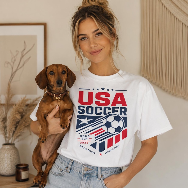 May include: White t-shirt with a USA Soccer graphic in red and blue, featuring a soccer ball design. The shirt also includes the text "World Cup 2026" and "Built on Passion".