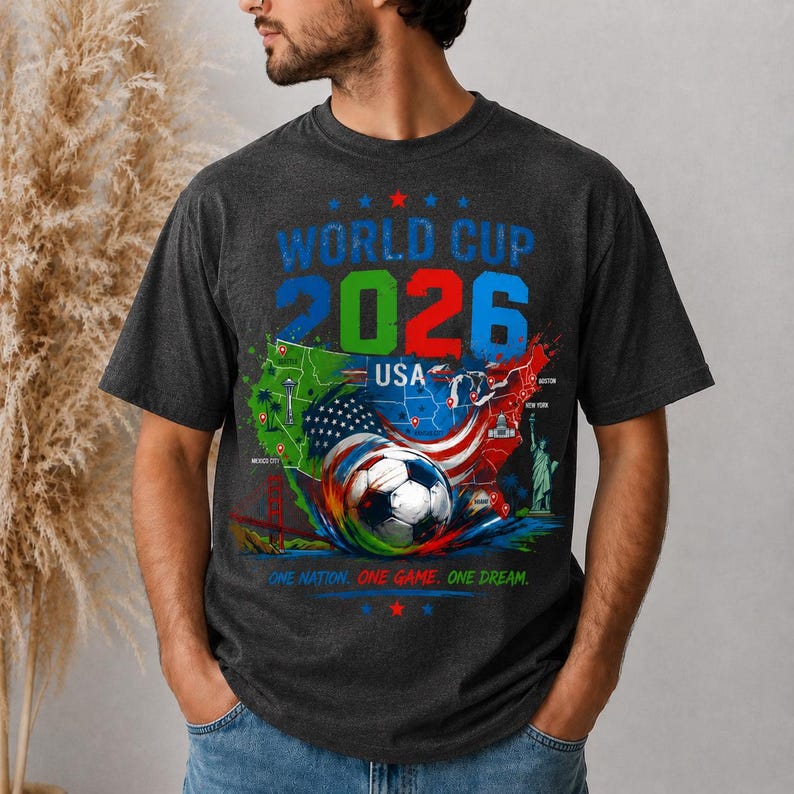 May include: Dark gray t-shirt featuring a colorful graphic for the 2026 World Cup USA. The design includes a soccer ball, a map of the USA, and the text "World Cup 2026 USA". The shirt also has the slogan "One Nation. One Game. One Dream."