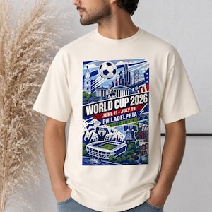 May include: A cream-colored short-sleeve t-shirt featuring a graphic print of the World Cup 2026 in Philadelphia. The design includes a soccer ball, city skyline, stadium, and the text World Cup 2026, June 11-July 19, Philadelphia, Host City.