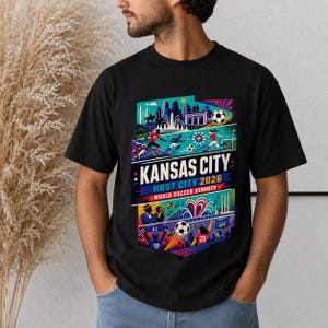 K&ouml;nnte beinhalten: Ein schwarzes T-Shirt mit einem farbenfrohen Grafikdruck und dem Text KANSAS CITY HOST CITY 2026 WORLD SOCCER SUMMER. Das Design zeigt Illustrationen von Fu&szlig;ballspielern, einer Skyline, einem Jazzmusiker und Fu&szlig;b&auml;llen in leuchtenden Farben.