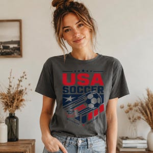 May include: Dark gray t-shirt with the text "USA SOCCER" in red and white. The design includes a soccer ball with a trail, a star, and the words "WORLD CUP" and "GAME OF SPORT". The shirt is made of a soft material.